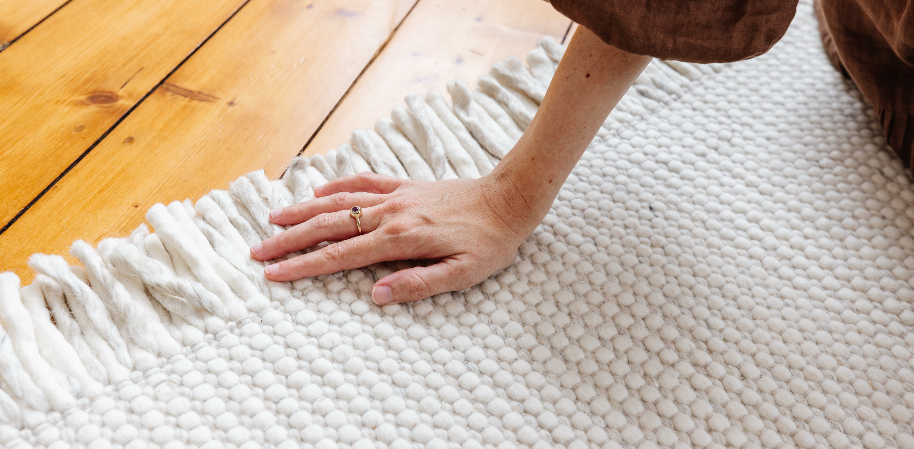 Ein heller Teppich von benuta liegt auf einem Holzboden, darauf sitzt eine Person. Im Bildausschnitt ist ihr Unterarm und ihre Hand zu sehen, die den Teppich berührt. Quelle: benuta