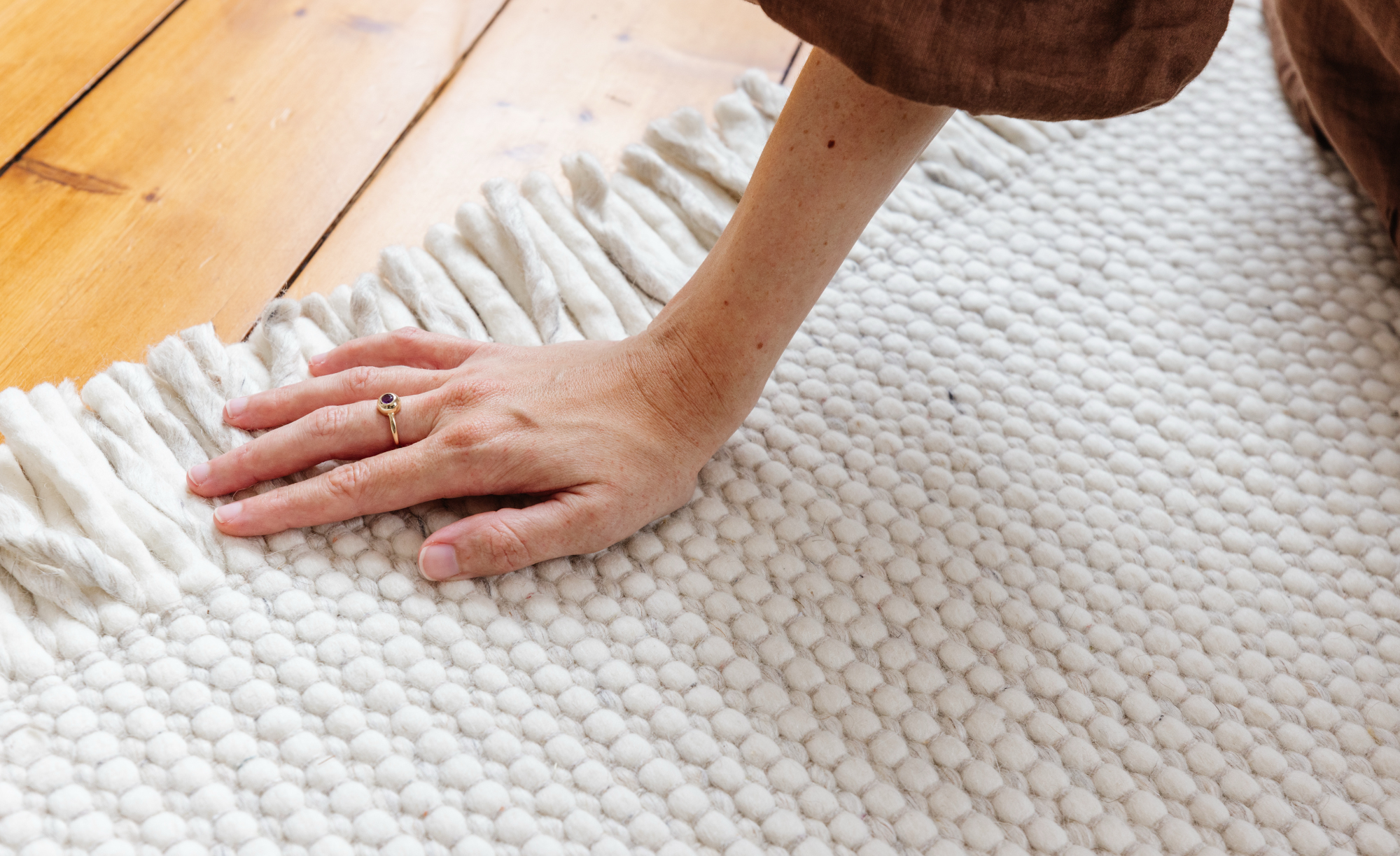 Ein heller Teppich von benuta liegt auf einem Holzboden, darauf sitzt eine Person. Im Bildausschnitt ist ihr Unterarm und ihre Hand zu sehen, die den Teppich berührt. Quelle: benuta